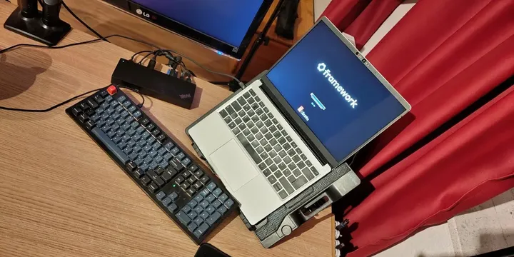 A top-down view of a wooden desk setup featuring a Framework laptop on a
stand, displaying the Framework logo and an Ubuntu loading screen. To the 
left is a mechanical keyboard with grey and black keycaps and a single red
accent key. The setup is connected to a ThinkPad docking station and a
secondary LG monitor. Red curtains are visible in the background.

